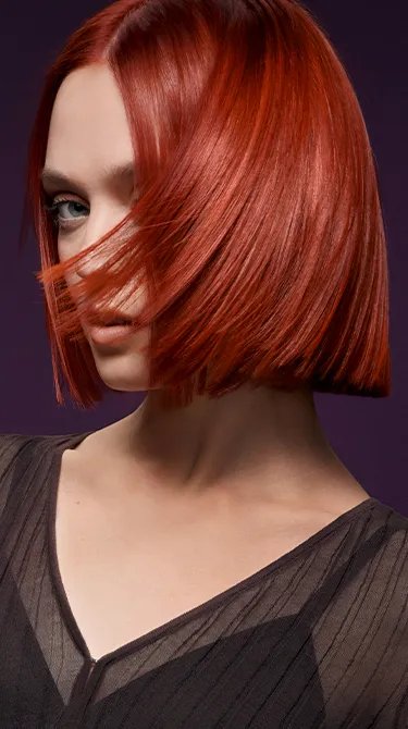 Image of model with a bright red, straight and short hairstyle against a dark purple background. Hair is slightly covering their face. 