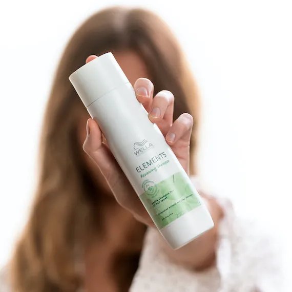 Woman holds Wella Professionals Elements Renewing Shampoo in front of her face. 