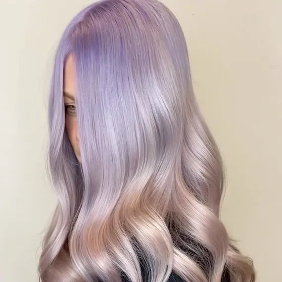 Close-up of long, wavy hair in a soft lavender shade with a gradient effect, transitioning from a deeper purple at the roots to a pastel tone towards the ends.