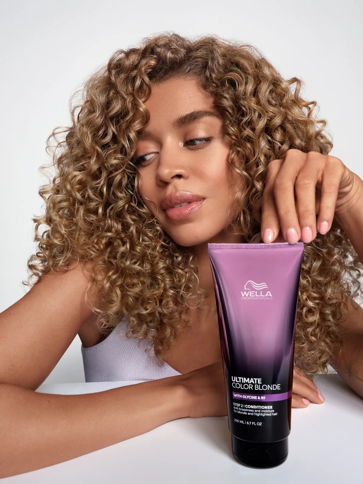 Model with caramel blonde, curly hair holds a bottle of ULTIMATE COLOR BLONDE Conditioner.