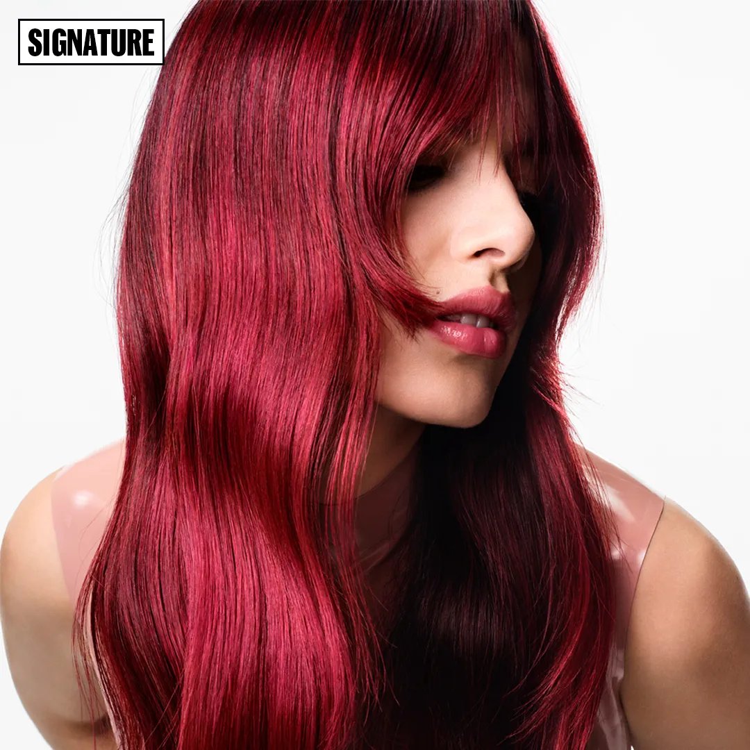 Profile view of a woman with long, straight, vibrant red hair with bangs, wearing a pale pink top against a white background.