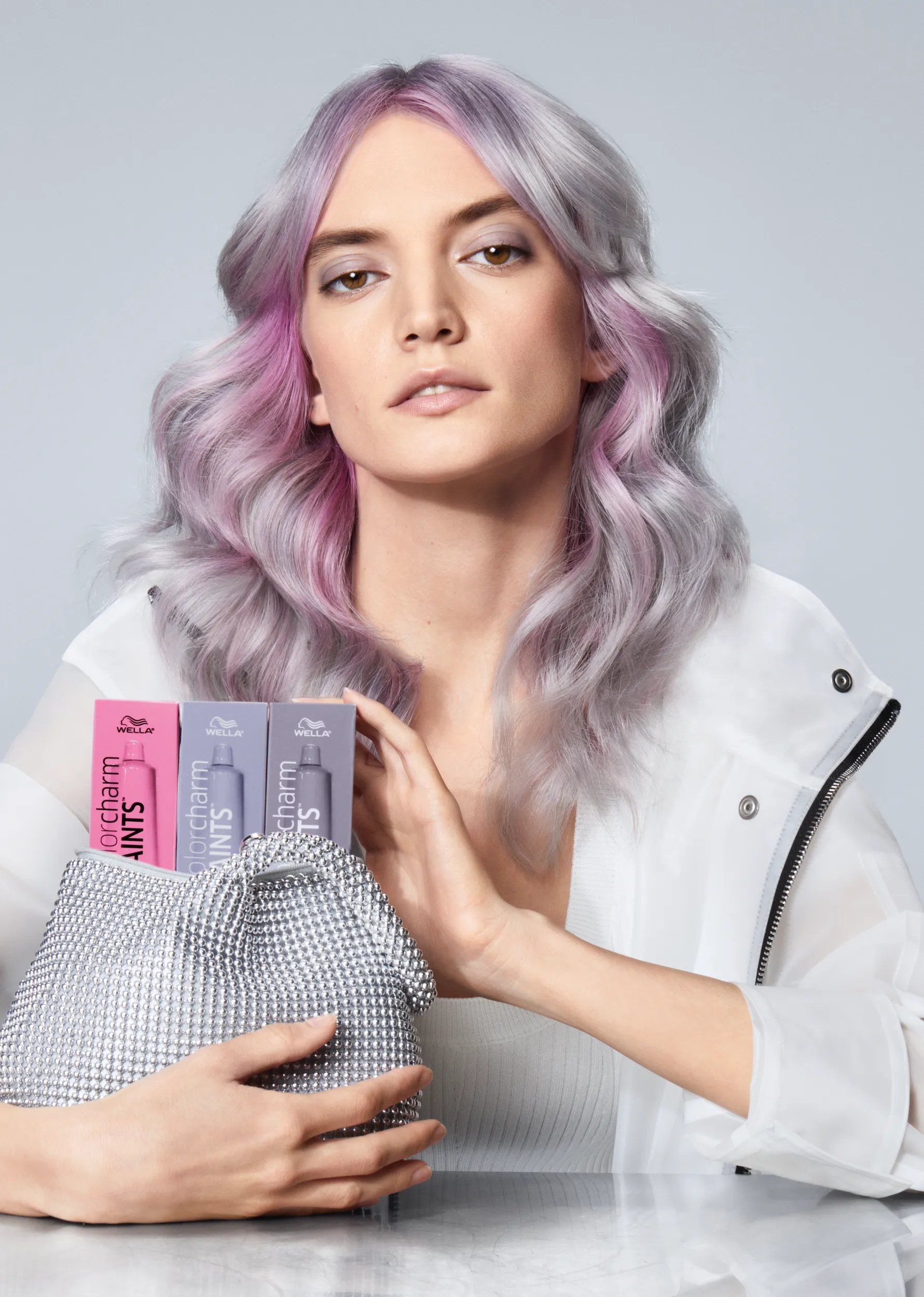 Model with silver and pink tinted wavy hair holding a silver sequin clutch with three Wella Colorcharm Paints boxes.