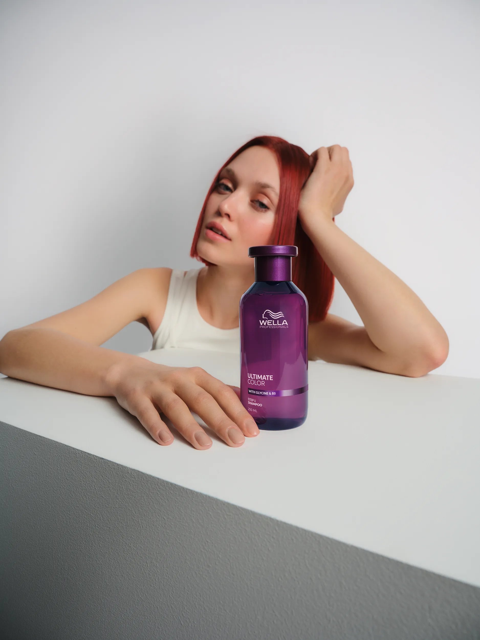 Image of model with curly brunette hair and red streaks, leaning their chin on one arm and holding the Ultimate Color conditioner with their other hand. Text overlay reads "Up to 86% less breakage* *vs non-conditioning shampoo". 			
