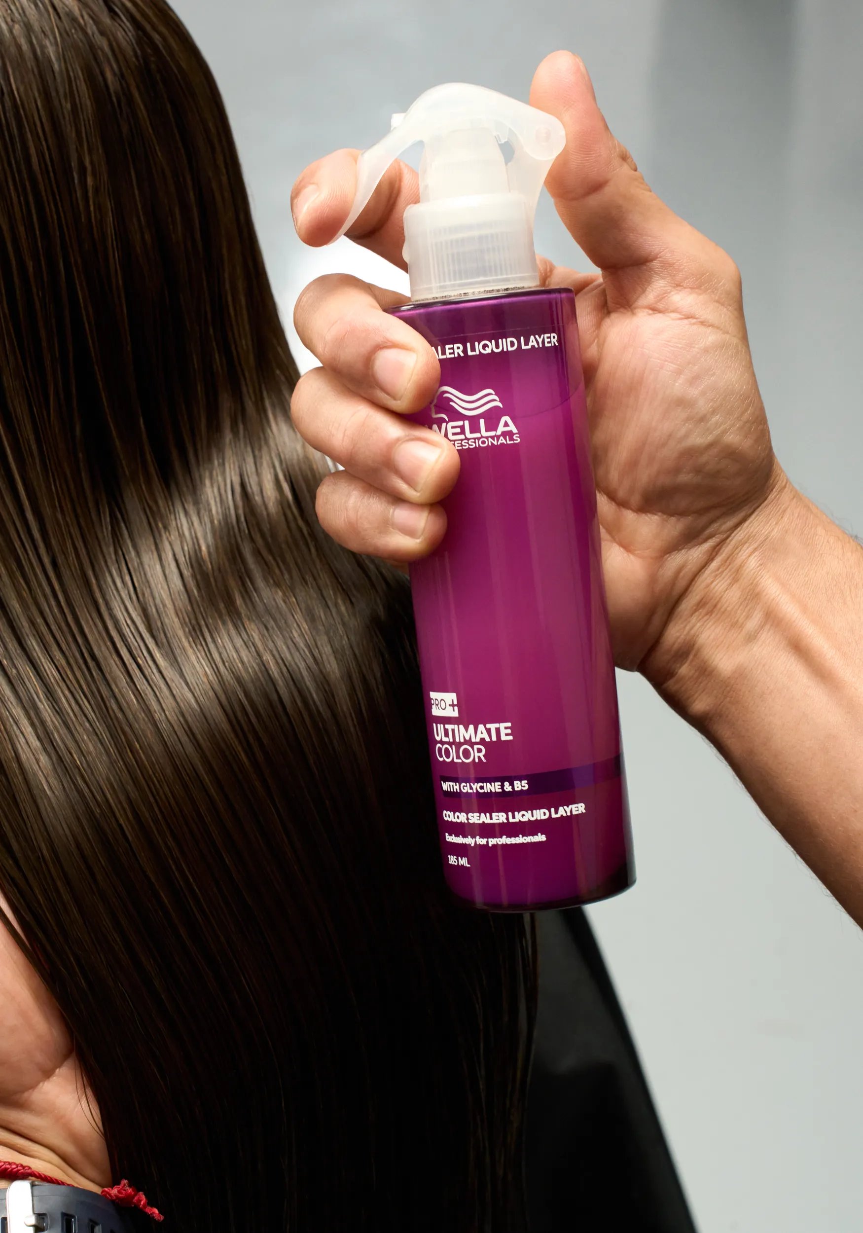 A side profile of a model with brunette curly hair and red highlights. Model is wearing a black top and is against a purple background. 