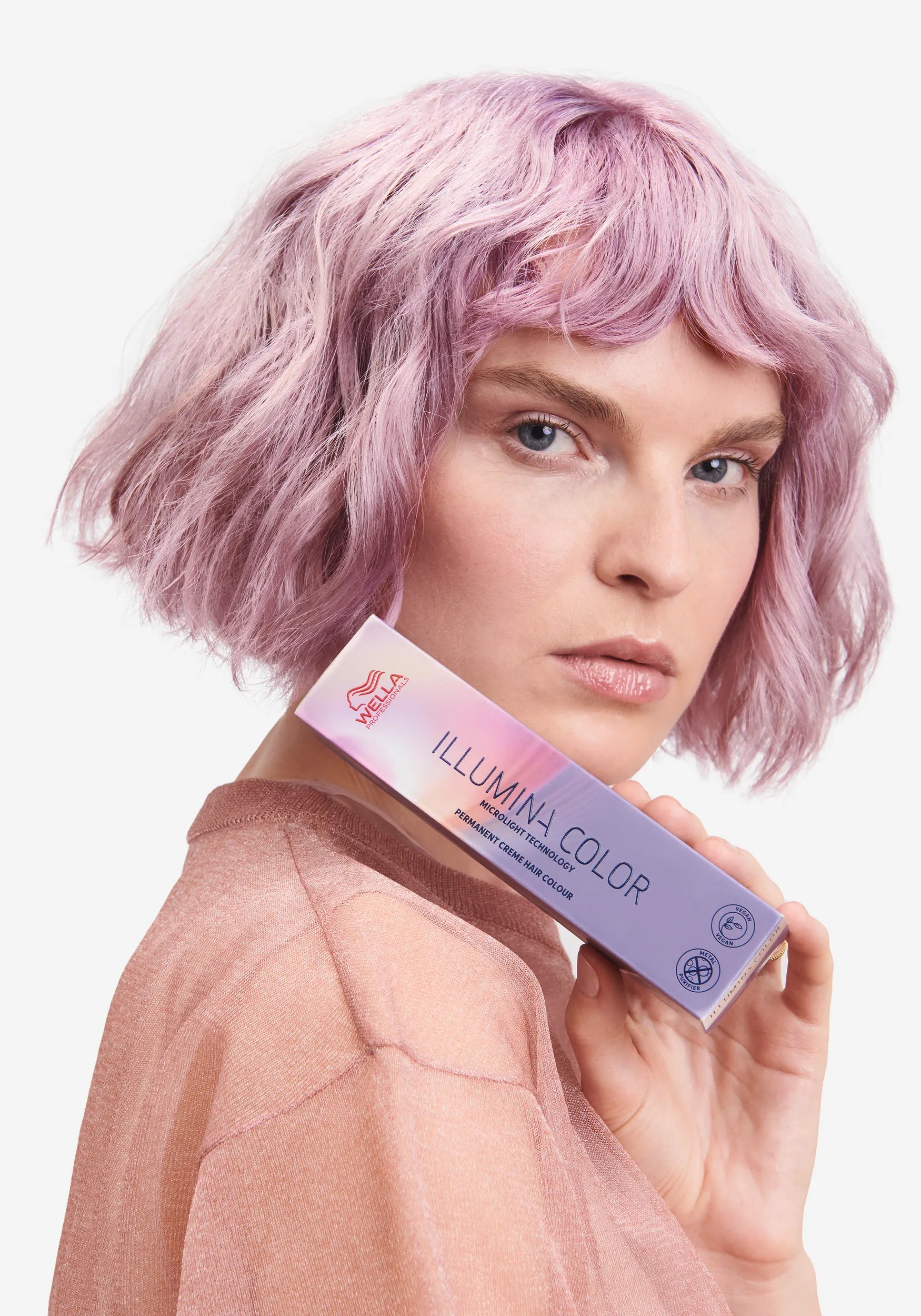 Woman standing sideways one with her head turned facing the camera. She has a light pink bob with a short fringe and is holding the Illumia color product to the camera.