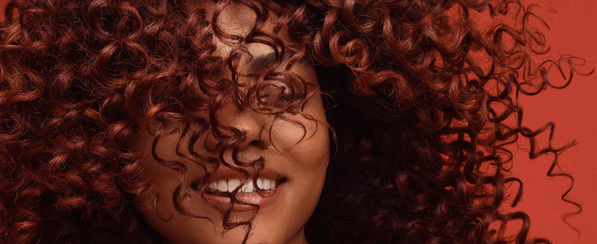 Close-up of a smiling woman with long, curly, auburn hair partially obscuring her face, set against a red background.