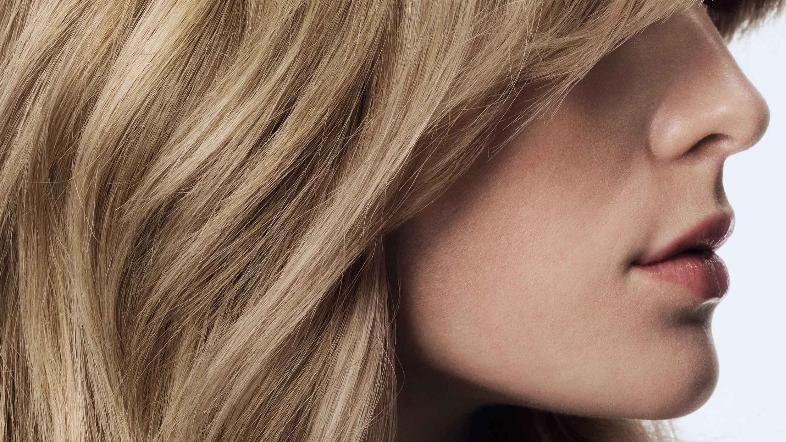 Side profile of a human face with wavy blonde hair, closed lips, and focus on the hair texture and facial outline against a neutral background.