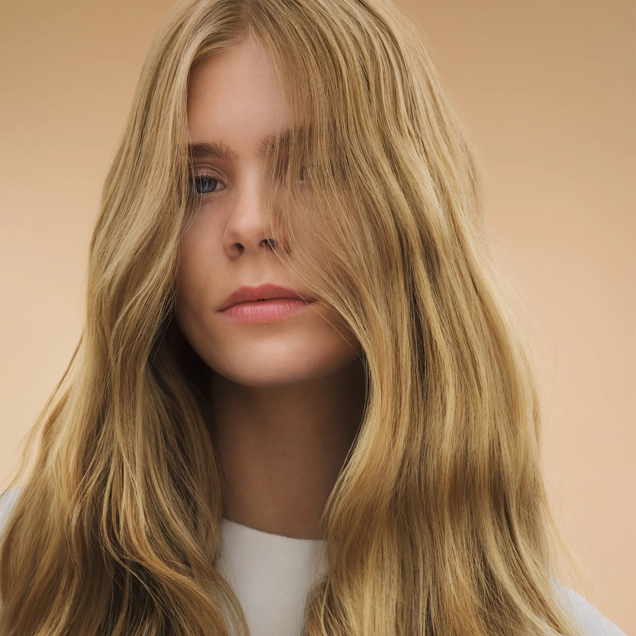 Porträt einer Person mit langem, wellig fallendem blondem Haar mit feinen Highlights vor neutralem Hintergrund.
