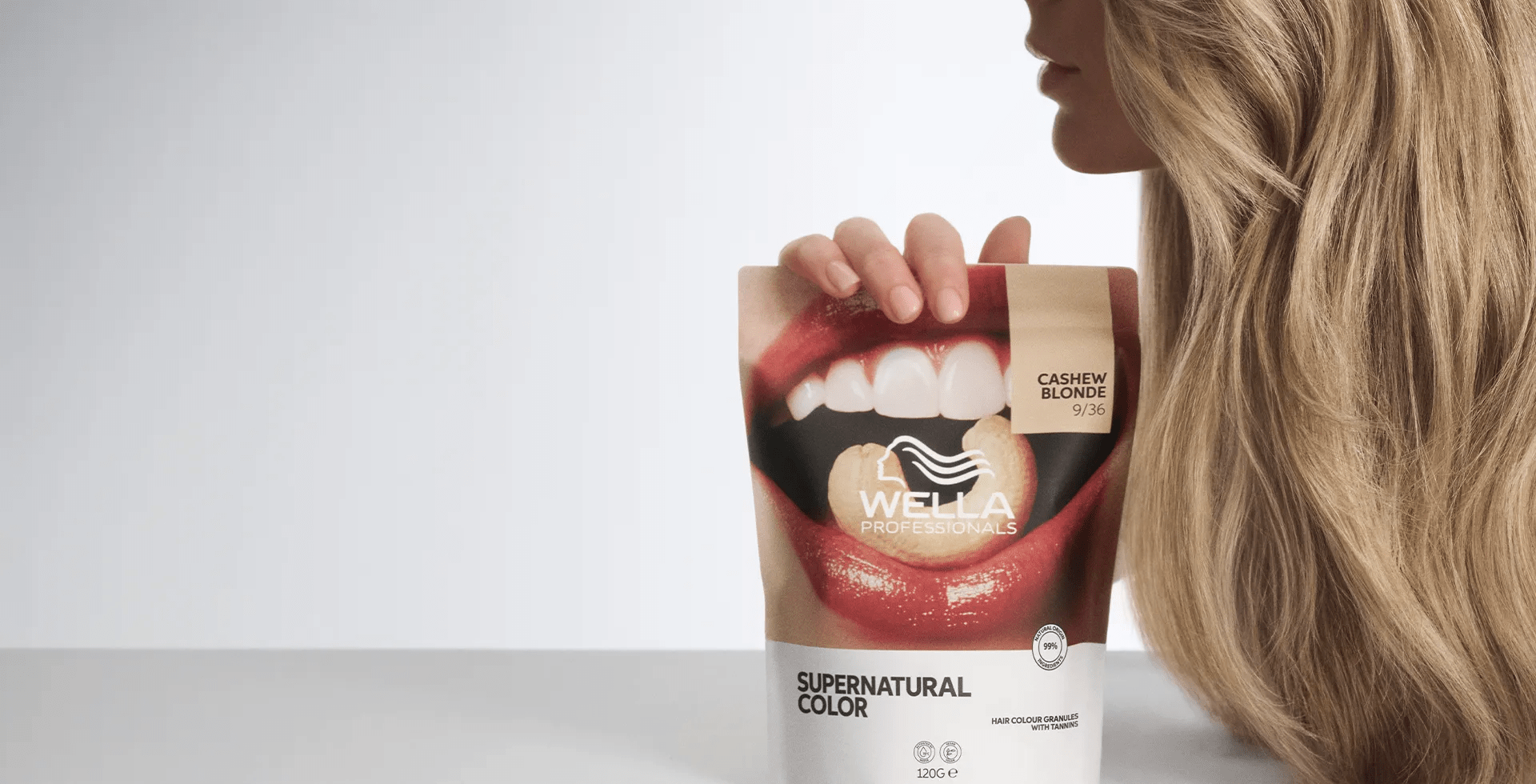 Εικόνα ενός μοντέλου σε προφίλ, που δείχνει τα ξανθά, κυματιστά μαλλιά του. Το μοντέλο κρατά στο χέρι του μια συσκευασία Wella Professionals Supernatural Color στην απόχρωση Cashew Blonde. Η εικόνα έχει τραβηχτεί σε λευκό φόντο.