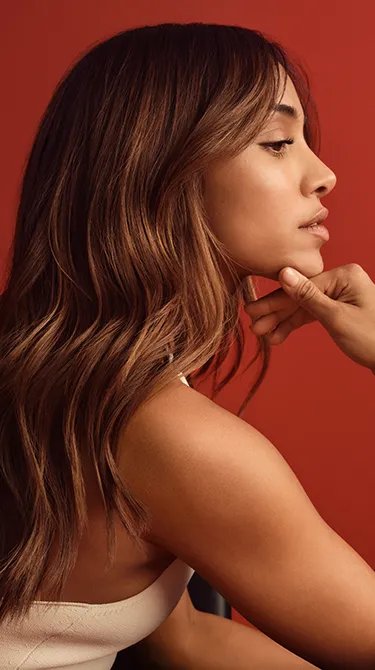 Profile portrait of a woman with wavy brown hair, resting her chin on her hand against a red background.