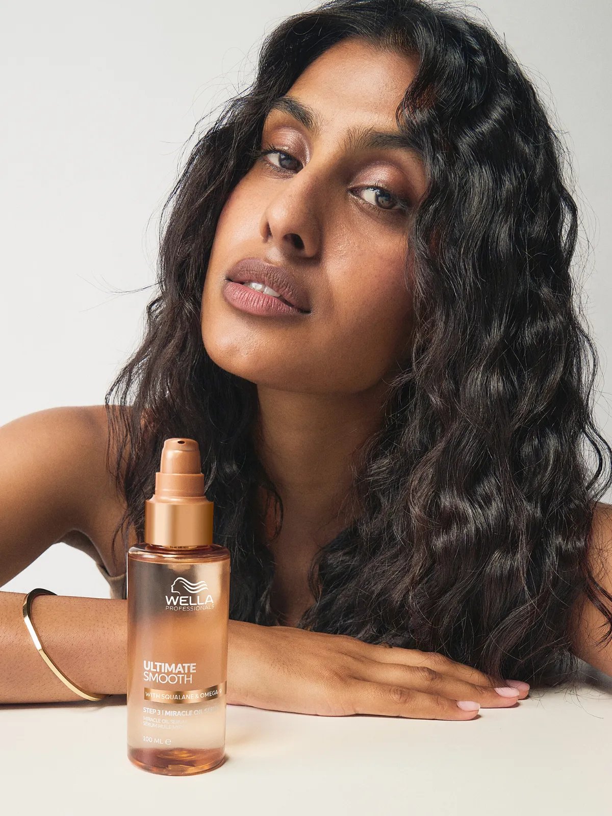 Model with dark brown, glossy, wavy hair poses next to a bottle of ULTIMATE SMOOTH Miracle Oil Serum. 