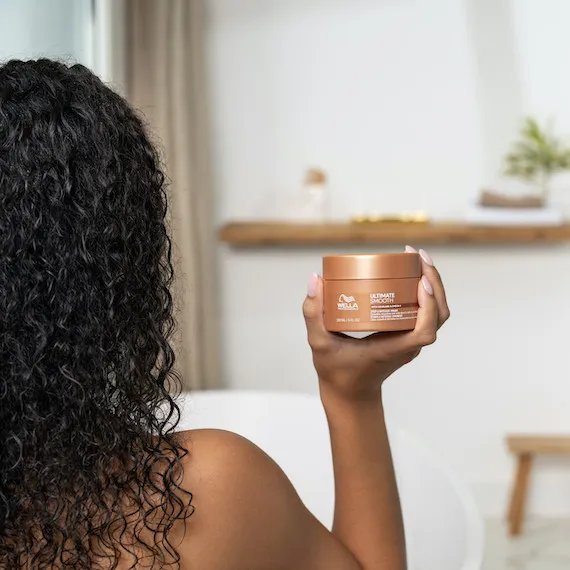 Model with dark brown, curly, wet hair holds up a bottle of ULTIMATE SMOOTH mask.