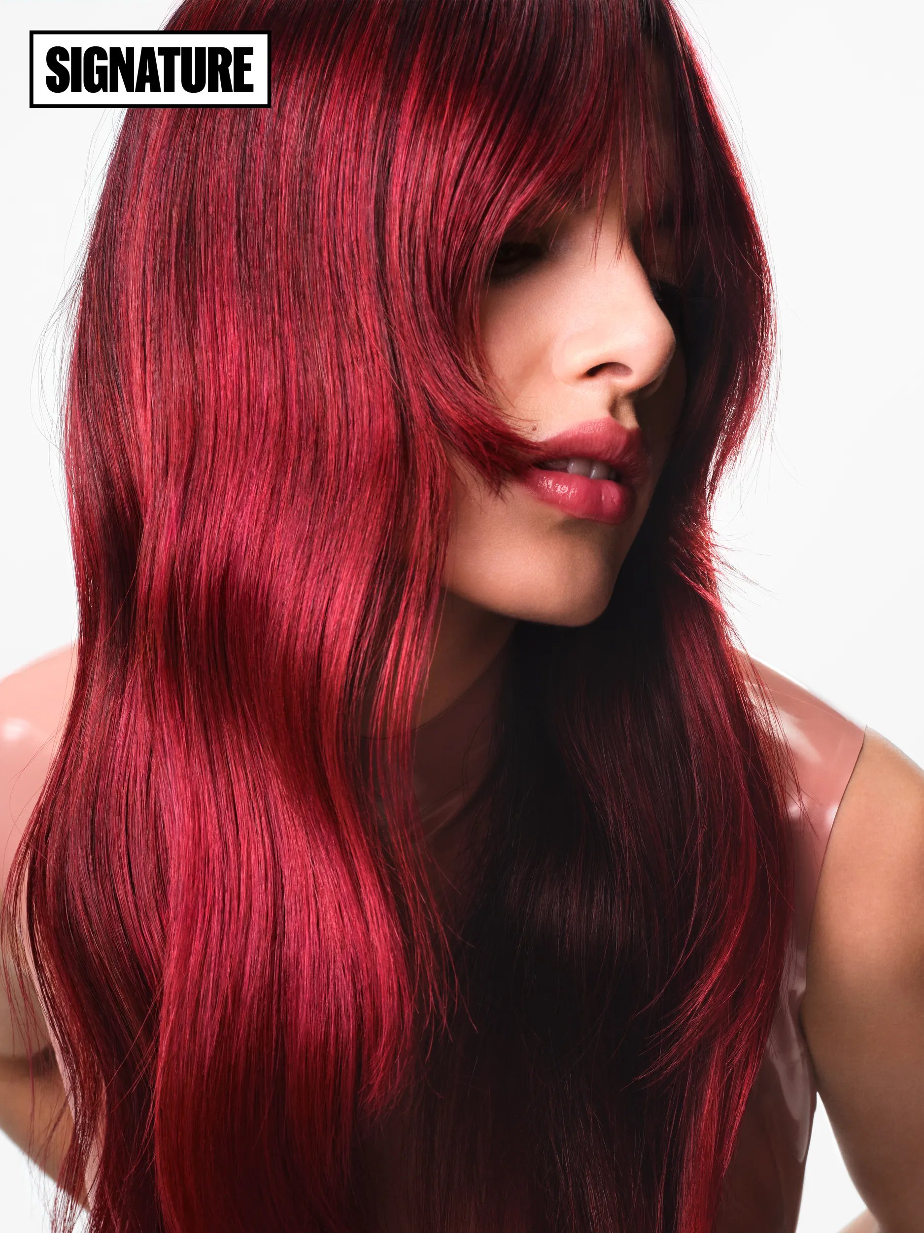 Close-up portrait of a woman with long, vibrant red hair styled with bangs, wearing a shiny pink top.